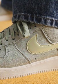 Grønne semskede Nike-sko med en blank grøn swoosh-logo, flade snørebånd og en struktureret hvid sål, delvist dækket af denim-bukser.