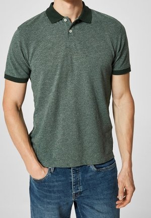 Man wearing a short-sleeve gray-green polo shirt with dark green collar and cuffs, paired with blue jeans, standing with one hand in pocket.