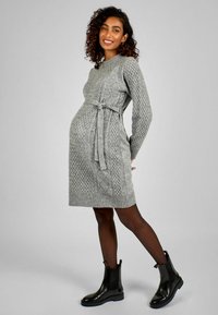 Vestito premaman grigio a maglia con motivi texturizzati, maniche lunghe e una cintura legata in vita. Indossato con stivaletti neri alla caviglia.