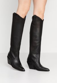 Czarne skórzane kozaki za kolano z ostro zakończonym noskiem i koturnem. Posiadają ozdobne przeszycia oraz pętelki u góry ułatwiające zakładanie.