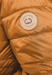 Brązowa kurtka puchowa z przeszytymi liniami; posiada okrągłą, beżową skórzaną łatę z napisem "Gerry Weber Outerwear" oraz emblematem.