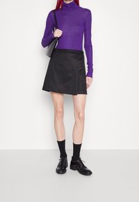 Lila långärmad polotröja, svartsnurrad kjol, svarta snörskor med spikar och svarta ankelsockor, mot en enkel bakgrund.