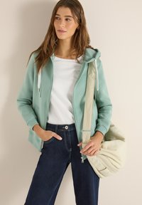 Femme portant un sweat à capuche zippé vert menthe, t-shirt blanc, jean foncé, tenant un sac à bandoulière beige, debout contre un mur uni.