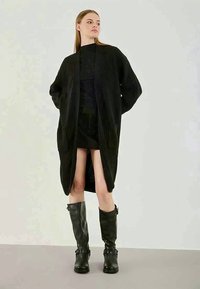 Nő, aki hosszú fekete bordás kardigánt, fekete ruhát és térd magas fekete bőrcipőt visel, háttal áll a kezével a háta mögött egy sima háttér előtt.