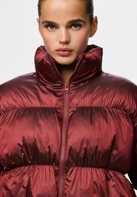 Burgundiškai raudonas pūkinis švarkas su aukštu apykakle, siuvinėtu paviršiumi ir centriniais užtrauktukais. Medžiaga atrodo blizgi ir lengva. Priekinis vaizdas.