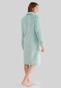 Mujer de pie, descalza, vistiendo un albornoz de felpa texturizada azul claro con los puños doblados, mirando ligeramente hacia un lado sobre un fondo gris liso.