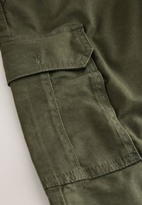 Olivovo zelená látka zobrazujúca detail cargo vrecka s chlopňou a kontrastným prešívaním. Hladká textúra, pevná konštrukcia.