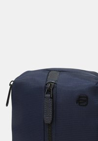 Primo piano di una trousse da viaggio testurizzata blu scuro con una cerniera nera centrale e un piccolo logo nero sul lato anteriore.