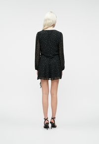 Kvinde med langt blondt hår iført en sort minikjole med hvide prikker og gennemsigtige lange ærmer, stående med ryggen til, i sorte stiletter.