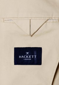 Tissu beige avec une étiquette rectangulaire bleu marine cousue, affichant le nom de la marque "Hackett London" et un logo avec des maillets de polo croisés.