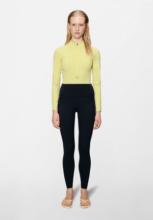 Γυναίκα που στέκεται με το πρόσωπο προς τα εμπρός, φορώντας ανοιχτό κίτρινο μακρυμάνικο μπλουζάκι με φερμουάρ, μαύρο κολάν με ψηλή μέση και μπεζ σαγιονάρες σε απλό φόντο.