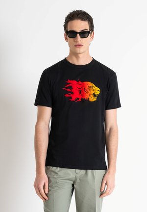 Zwarte katoenen t-shirt met een kleurrafbeelding van een leeuw in oranje en rood met vlammen. Gecombineerd met lichtgroene broek. Eenvoudige ronde halslijn.