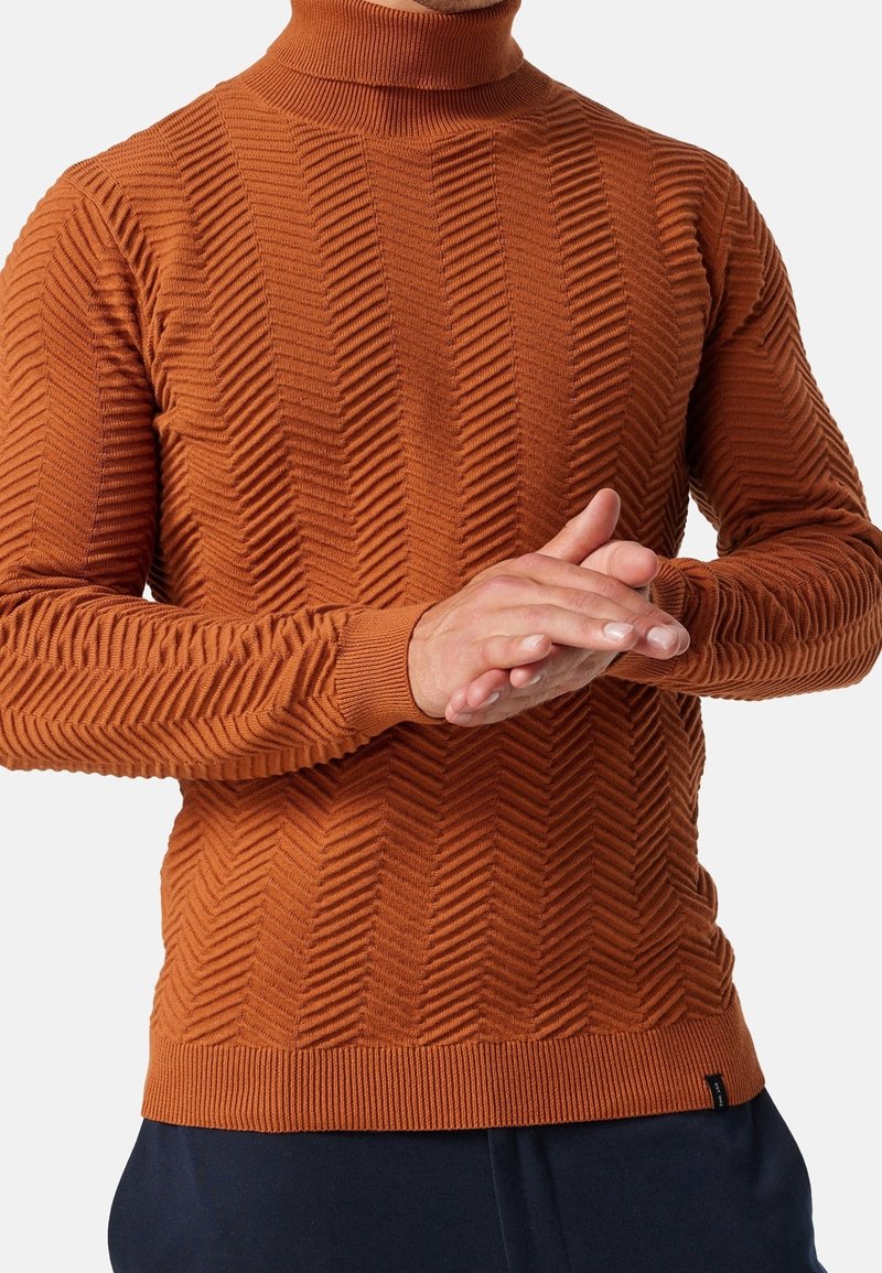 Homme portant un pull à col roulé en tricot texturé orange rouille avec des motifs en chevrons, joignant les mains devant lui sur un fond blanc.