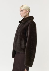 Femme blonde de profil portant une veste en fausse fourrure marron foncé, des boucles d'oreilles anneaux dorées et une jupe côtelée sombre avec une fente à l'avant.