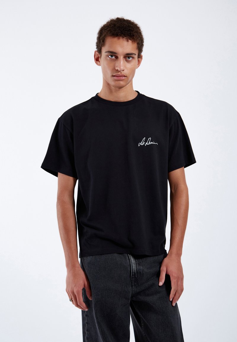 T-shirt in cotone nero con vestibilità rilassata, scollatura a girocollo e dettaglio ricamato in bianco con firma sul petto. Abbinato a pantaloni in denim scuro.