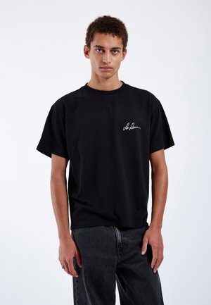 Black cotton t-shirt featuring short sleeves and a round neckline, with a white signature logo on the left chest. Worn with dark jeans.
