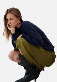 Granatowy sweter z prążkowanymi mankietami, oliwkowa plisowana spódnica i czarne kozaki do kolan. Zawiera złote wyraziste kolczyki.