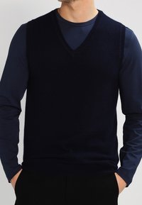 Mörkblå V-ringad tröja, tillverkad av mjukt material. Ärmdesign utan ärmar med en slät textur och fin avslutning. Bärs över en långärmad topp.