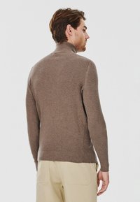 Brązowy, teksturowany sweter dzianinowy z wysokim kołnierzem, dopasowanym krojem i długimi rękawami, w połączeniu z jasnymi spodniami.