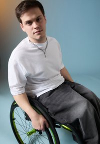 Osoba w białej koszuli z krótkim rękawem i szarych spodniach siedzi na czarno-zielonym wózku inwalidzkim na tle jasnoniebieskim.