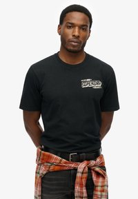 T-shirt di cotone nero a maniche corte, con un logo stampato bianco sul petto. Indossato con una camicia di flanella a quadri arancione legata in vita.