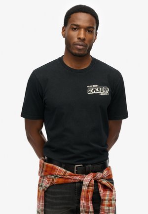 T-shirt noir en coton à manches courtes, avec un logo imprimé blanc sur la poitrine. Porté avec une flanelle orange à carreaux nouée à la taille.