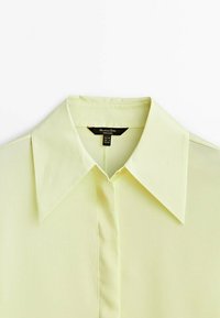 Camicia collare gialla chiara realizzata in tessuto liscio; presenta una chiusura a bottoni e un design classico del colletto, con un'etichetta visibile all'interno.