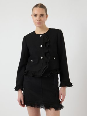 Svart ensemble med en ruffle-jakke med sølvknapper og matchende skjørt, begge med gjennomskinnelige ruffle-detaljer i kantene.