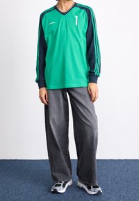 Henkilö, jolla on päällään vihreä ja tummansininen pitkähihaisen Adidas-paita, jossa on numero 1, löysät harmaat farkut ja valkoiset, mustat ja siniset lenkkarit.