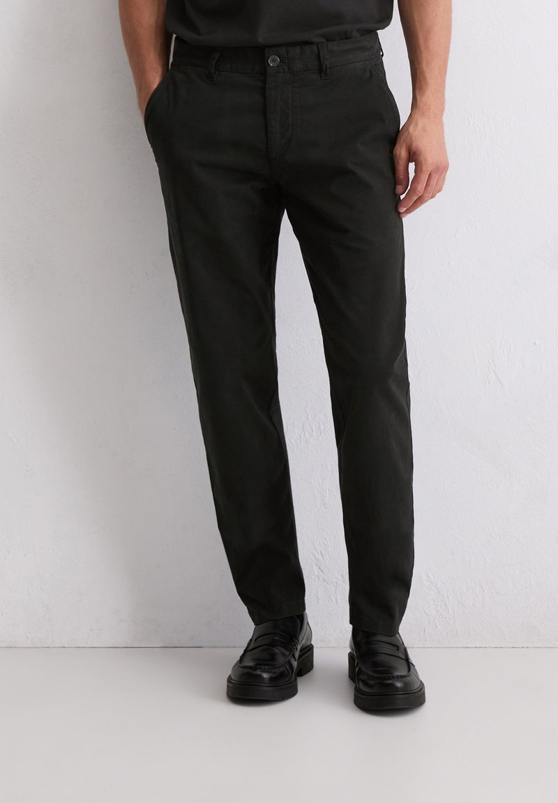 Pantalon en coton noir avec une coupe droite, doté d'une fermeture à bouton et de poches latérales, associé à des chaussures noires.
