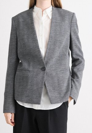 Person iført en grå sildebensblazer over en hvid skjorte med knapper og sorte bukser mod en ensfarvet baggrund.