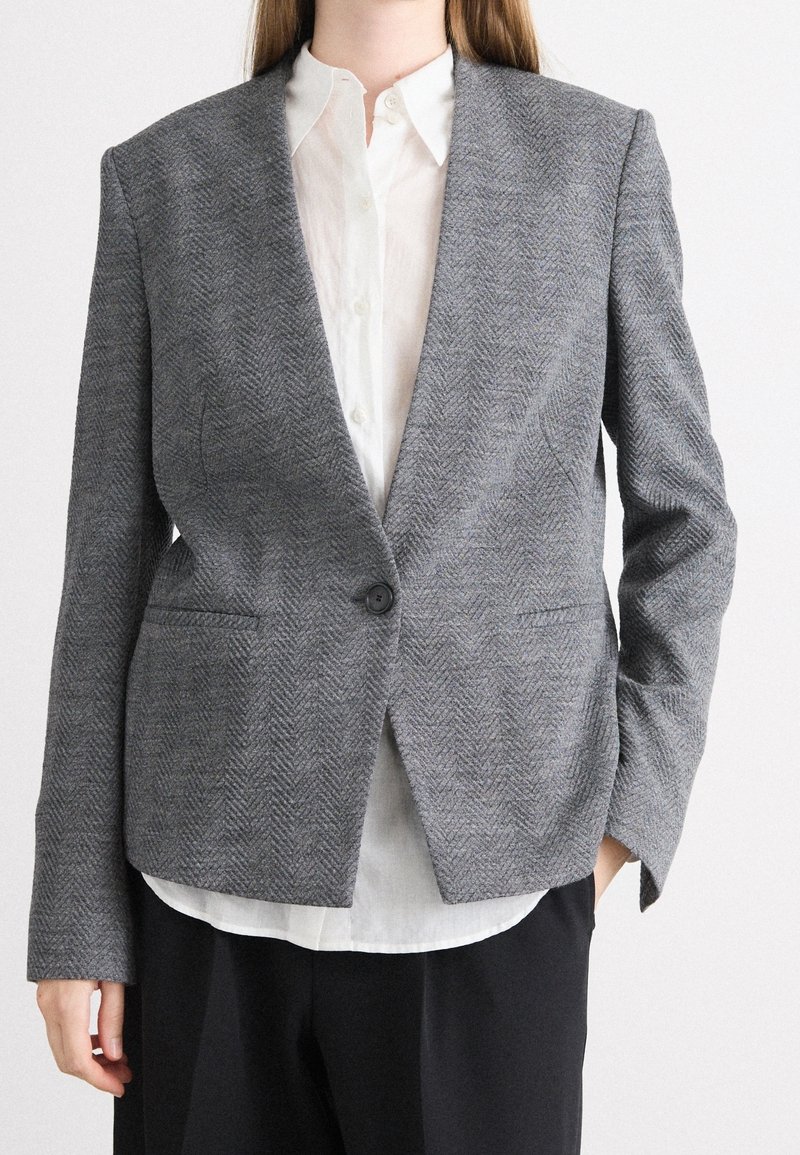 Personne portant un blazer en chevrons gris sur une chemise blanche boutonnée avec un pantalon noir sur un fond uni.