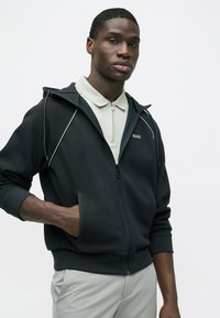 Μαύρο φούτερ με φερμουάρ και λευκές λεπτομέρειες, κατασκευασμένο από υφή υφάσματος. Το μοντέλο φοράει ανοιχτό γκρι πόλο από μέσα, με μικρό γιακά.