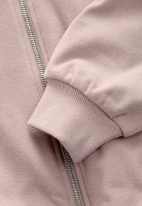 Lichtroze zip-up jack met ribgebreide manchetten en ritsdetails, gemaakt van zachte, gestructureerde stof met een gladde afwerking.