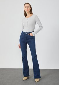 Sivý dlhý rukáv henley top s gombíkmi, skombinovaný s tmavými riflovými širokými džínsami a béžovými lodičkami na vysokom opätku na neutrálnej pozadí.