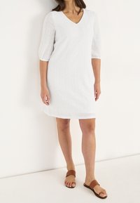 Vit, kortärmad klänning med V-ringning, har ögla detaljer och en avslappnad passform. Kombinerad med platta bruna sandaler.