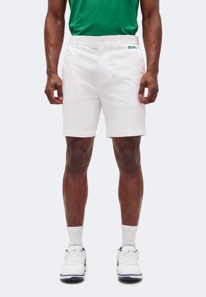 Άνδρας που φοράει λευκά, ραμμένα σορτς με κουμπί και φερμουάρ, σε συνδυασμό με λευκά αθλητικά παπούτσια, κάλτσες και πράσινο πουκάμισο μερικώς ορατό.