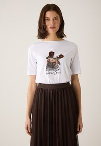 Weißes T-Shirt aus Baumwolle mit einem Grafikdesign eines Pugs, der Geweih und einen Schal trägt, kombiniert mit einem braunen Faltenrock. Hat kurze Ärmel und einen runden Halsausschnitt.