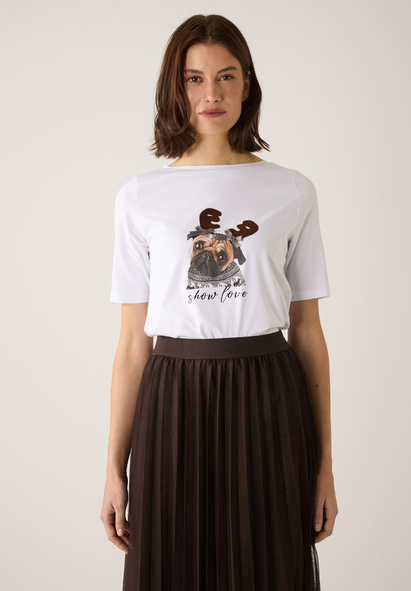 Weißes T-Shirt aus Baumwolle mit einem Grafikdesign eines Pugs, der Geweih und einen Schal trägt, kombiniert mit einem braunen Faltenrock. Hat kurze Ärmel und einen runden Halsausschnitt.