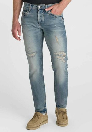 Mann trägt hellblaue, zerrissene Jeans und beigefarbene Wildleder-Schnürschuhe, steht vor einem einfarbigen Hintergrund.