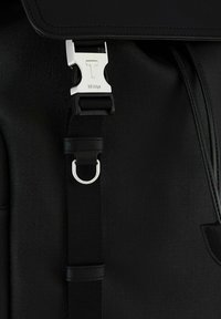 Schwarz strukturierter Rucksack mit einer silbernen Metallschnalle mit einem 'T', einem schwarzen verstellbaren Gurt und einem runden silbernen Ring für zusätzliche Unterstützung.