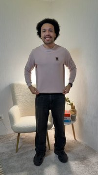 Man met krullend haar staat glimlachend in een beige shirt met lange mouwen en een zwarte broek in een zacht verlichte kamer met een witte, getextureerde fauteuil.