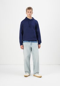 Hoodie blu navy con tasca frontale e cappuccio con cordino, abbinato a jeans blu chiaro e sneakers bianche. Tessuto morbido, vestibilità rilassata.