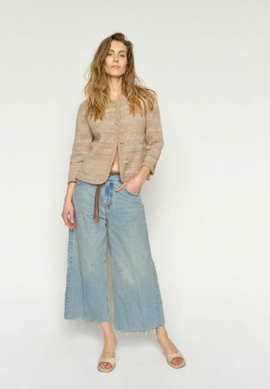 Naine seisab valgel taustal, kandes beežikat kootud kardigani, laia säärega lühikesi helesiniseid teksaseid pruuni vööga ning beeže avatud varbaga sandaale.