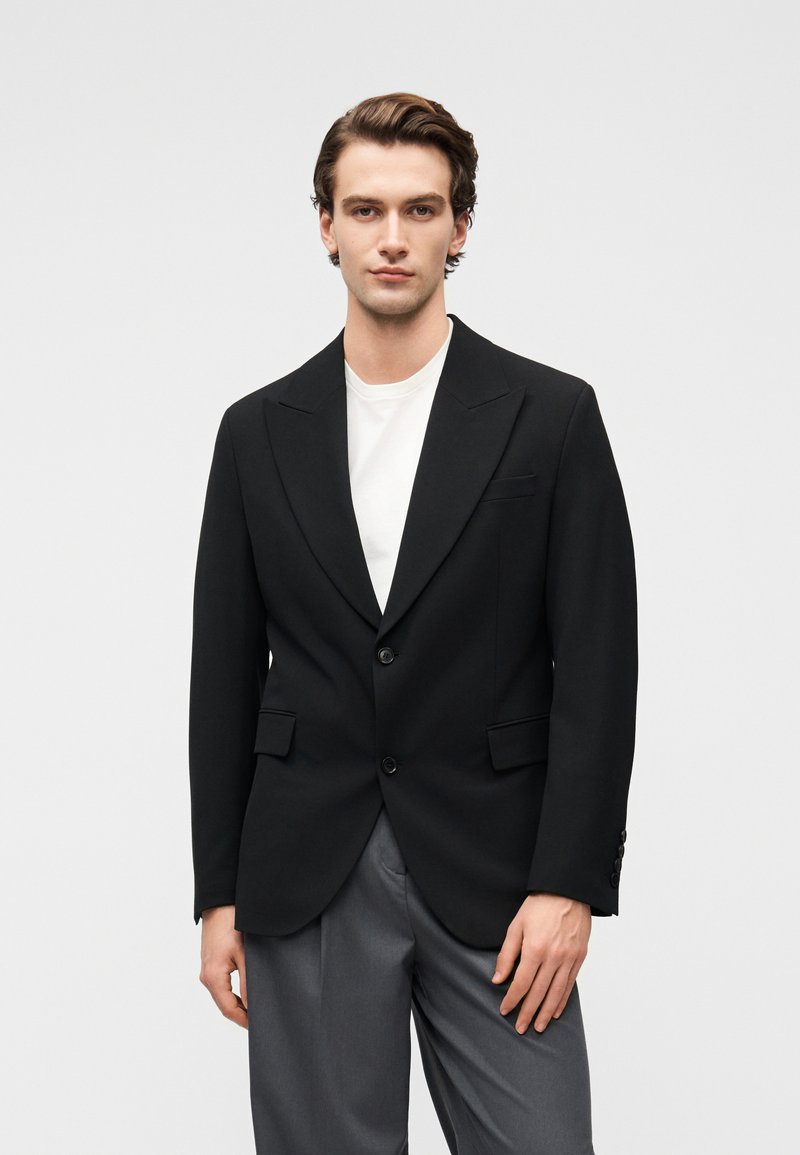 Sort blazer lavet af tekstureret stof, med en sporet revers, to knapper og forlommer, båret over en hvid t-shirt og grå bukser.