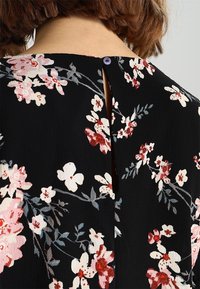 Tissu noir à motif floral, comprenant des fleurs roses et rouges. Un petit bouton violet se trouve à l'arrière, près d'une ouverture en forme de goutte d'eau.