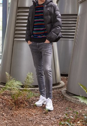 Chaqueta de plumón negra, suéter de rayas multicolores, jeans ajustados grises y zapatillas blancas. El fondo presenta estructuras metálicas y vegetación.