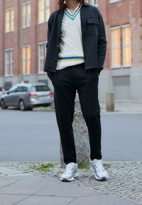 Svart jacka med ficka, krämfärgad kabelstickad tröja med blå och gula ränder, svarta byxor och silverfärgade sneakers, står på en trottoar.