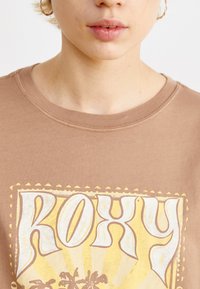 Bruine katoenen t-shirt met gele en witte grafische ontwerpelementen, inclusief het woord "ROXY" en een zonnestraalpatroon, ronde halslijn en korte mouwen.