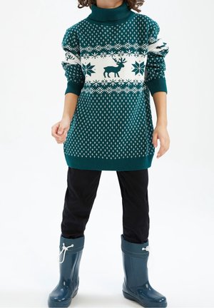 Dziecko ubrane w zielony sweter w renifery, czarne spodnie i turkusowe kalosze na białym tle.
